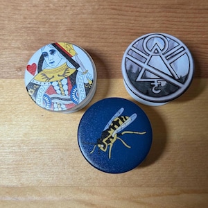 Könnte beinhalten: Drei runde PopSockets mit verschiedenen Designs. Eines zeigt die Herzkönigin Spielkarte, ein anderes ein geometrisches Design und das dritte eine gelb-schwarze Wespe auf marineblauem Hintergrund.