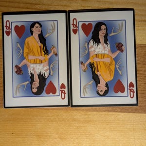 May include: Two playing cards featuring a woman with long dark hair and a yellow shawl. The cards are the Queen of Hearts. The woman is holding antlers and a heart in each hand. The background is blue.