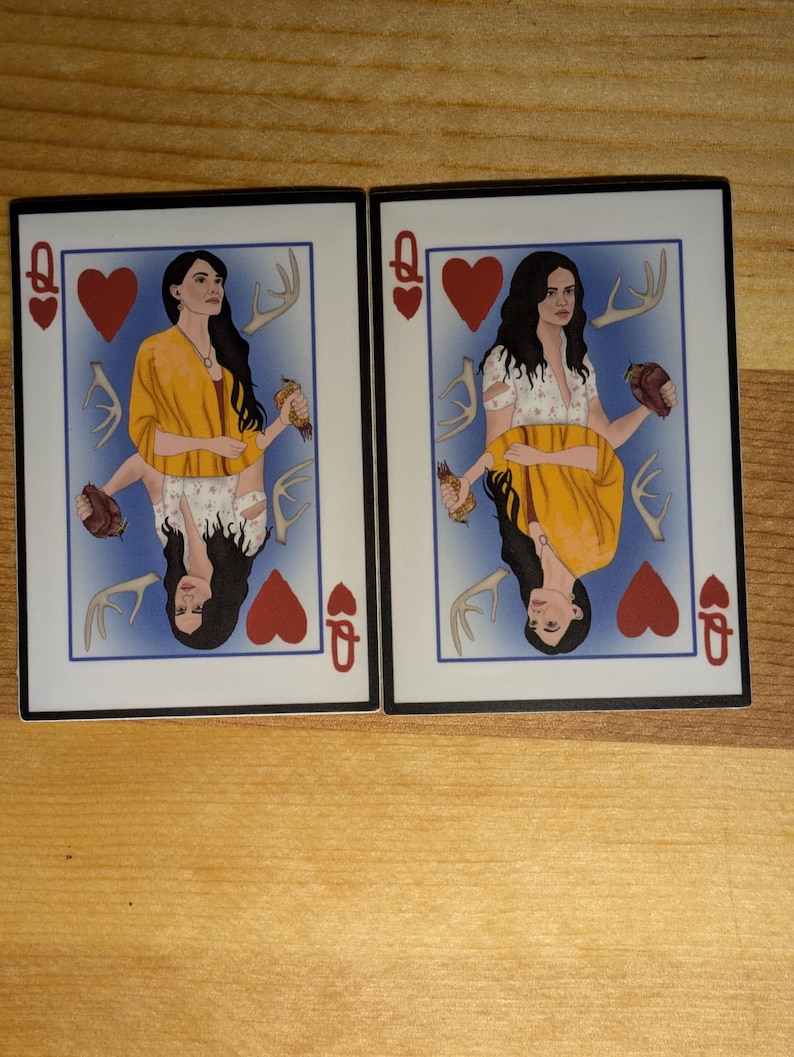 May include: Two playing cards featuring a woman with long dark hair and a yellow shawl. The cards are the Queen of Hearts. The woman is holding antlers and a heart in each hand. The background is blue.