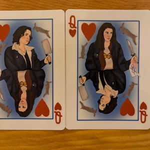 May include: Two playing cards featuring a queen of hearts with a woman holding a knife and a butcher knife. The cards are white with red hearts and a blue background.