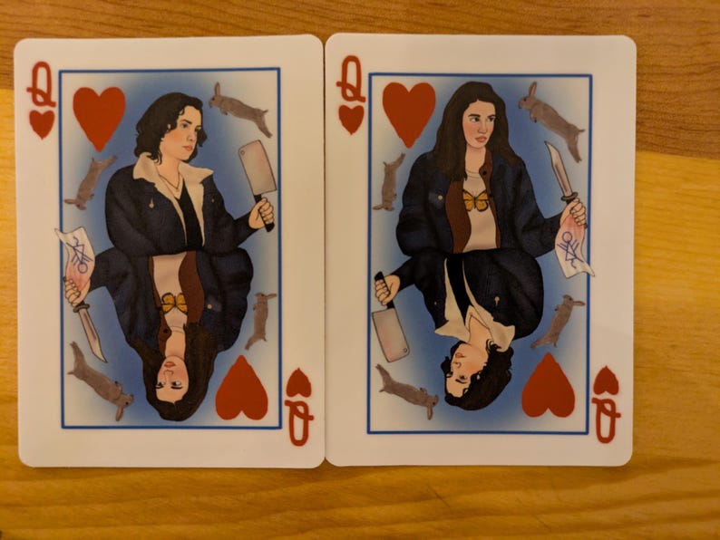 May include: Two playing cards featuring a queen of hearts with a woman holding a knife and a butcher knife. The cards are white with red hearts and a blue background.