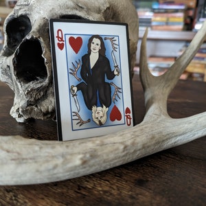 May include: A playing card with a queen of hearts design. The queen is depicted as a woman with dark hair and a black dress, holding a knife. The card is on a wooden surface with a skull and antlers.
