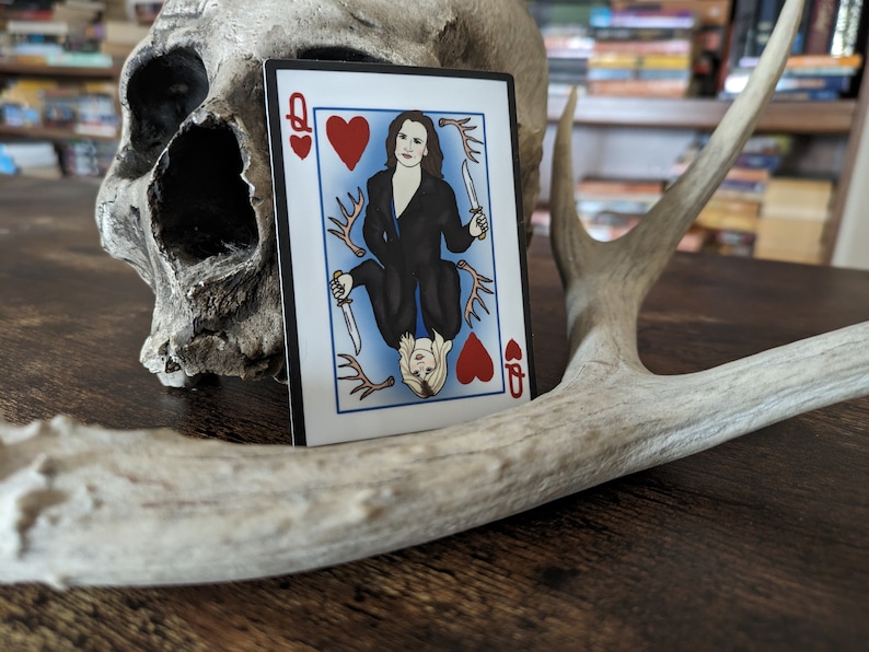 May include: A playing card with a queen of hearts design. The queen is depicted as a woman with dark hair and a black dress, holding a knife. The card is on a wooden surface with a skull and antlers.