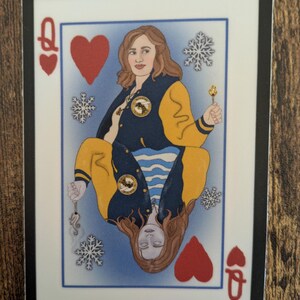 May include: A playing card featuring a woman with long brown hair wearing a yellow and blue jacket. The card is a Queen of Hearts and has snowflakes and a lit match on a blue background.