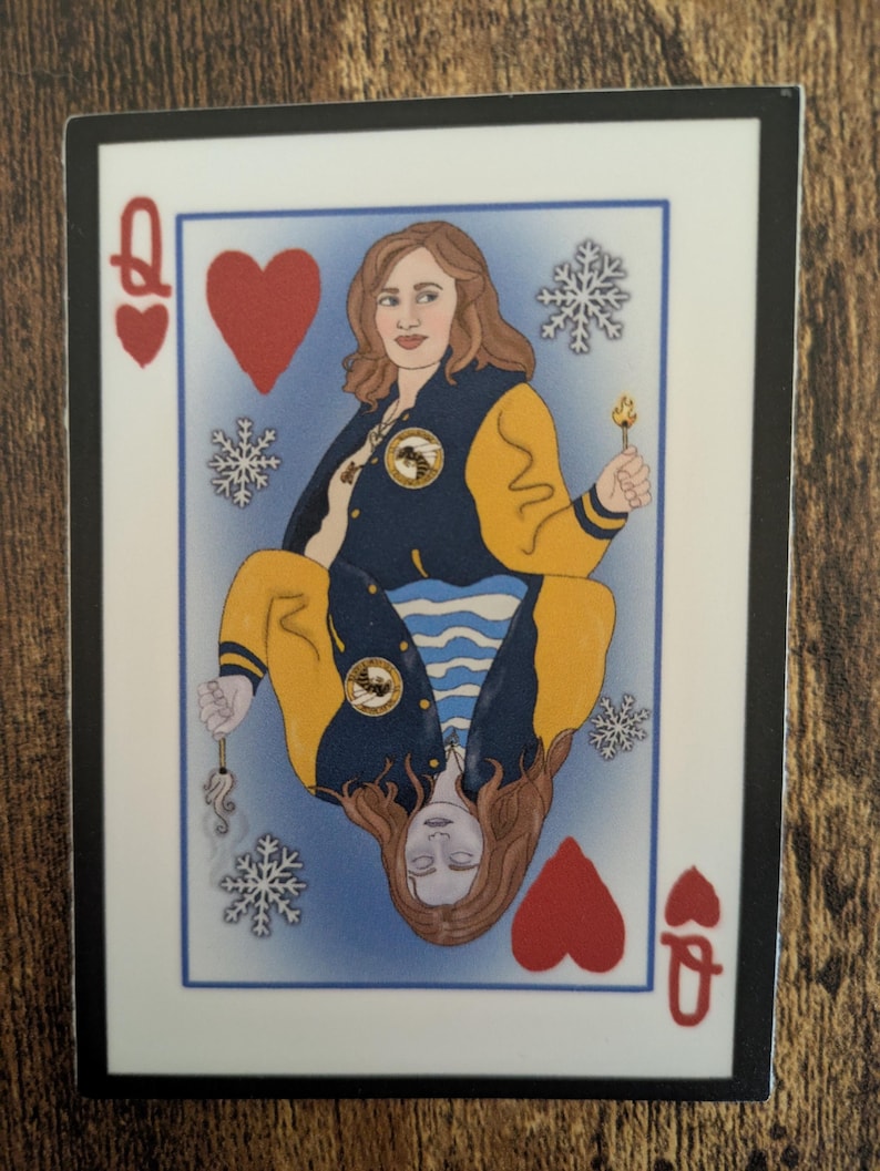 May include: A playing card featuring a woman with long brown hair wearing a yellow and blue jacket. The card is a Queen of Hearts and has snowflakes and a lit match on a blue background.