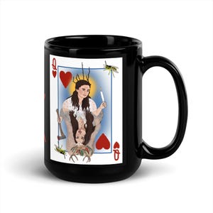 Puede incluir: Taza de cerámica negra con un diseño de carta de juego. La carta presenta a una mujer sosteniendo una vela y un hacha, enmarcada por un borde blanco, corazones rojos y la letra "Q". La taza tiene un acabado brillante.