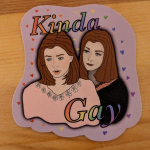 May include: A sticker with a purple background and rainbow text that says "Kinda Gay". Two women with brown hair are pictured on the sticker. The woman on the left is wearing a pink shirt with white flowers and the woman on the right is wearing a black shirt. The sticker is surrounded by small hearts.