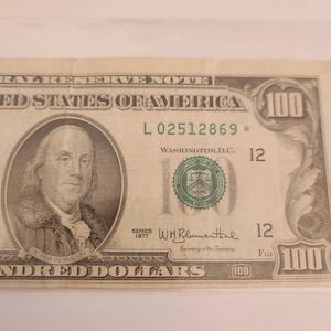May include: A United States one hundred dollar bill featuring a portrait of Benjamin Franklin. The bill includes the text "The United States of America" and "One Hundred Dollars". The bill is from Series 1977.