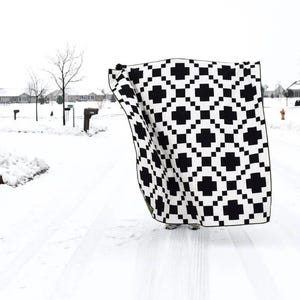 May include: A black and white geometric patterned blanket. The blanket features a repeating pattern of black plus signs and squares on a white background. The blanket is being held up in a snowy outdoor setting.