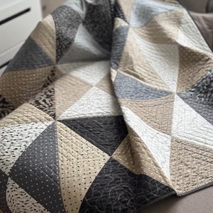 May include: A patchwork quilt with a geometric design. The quilt features beige, grey, and black triangular patterns. The quilt is made from various textured fabrics. It is likely used for bedding or decoration, and is draped over a sofa.