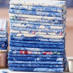 Puede incluir: Una pila de telas plegadas en varios tonos de azul y blanco, con diseños florales. Las telas están cuidadosamente apiladas, mostrando diversos patrones, incluyendo rosas y otros motivos botánicos. Se ven un carrete de hilo y un botón.