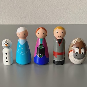 May include: A set of five wooden peg dolls featuring characters from a popular animated film. The dolls include a snowman, two female characters in blue and purple dresses, a male character in gray, and a painted rock resembling a reindeer.