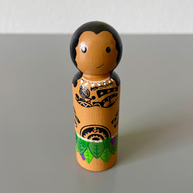 May include: A wooden peg doll with a tan body, black hair, and a smiling face. The doll is adorned with black tribal tattoos on the chest and torso, and a green and purple skirt. The doll is a decorative item.