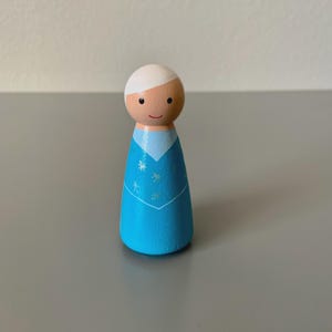 May include: A wooden peg doll featuring a light blue dress with snowflake details and a white collar. The doll has a round, wooden head with a white hair and a smiling face. This handcrafted doll is a charming decorative item.