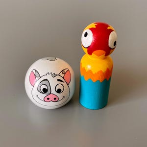 May include: Two hand-painted wooden toys: a white egg-shaped figure with a pig face and a peg doll parrot with a red head, yellow chest, orange accents, and blue body. The toys are designed with cartoon-like features and bright colors.