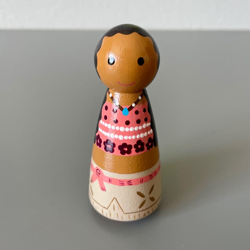 May include: A hand-painted wooden peg doll with a brown complexion, wearing a pink top with black polka dots and floral accents. The doll has a white skirt with a pink ribbon and gold accents. A blue gem necklace is also visible.