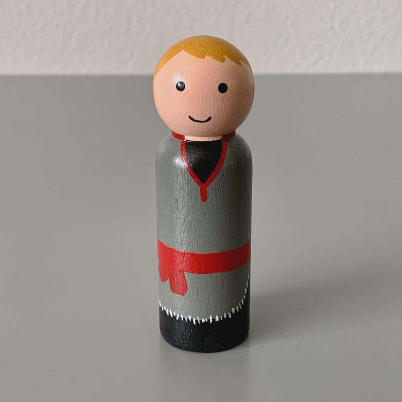 May include: A wooden peg doll with a painted design. The doll has a light skin tone, blonde hair, and a smiling face. It wears a gray tunic with a red belt and black pants. The tunic has a black collar.