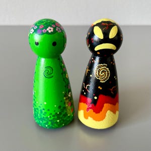 May include: Two hand-painted wooden peg dolls. One is green with floral details and a spiral symbol. The other is black with a yellow face, a spiral design, and a gradient of red, orange, and yellow at the base.