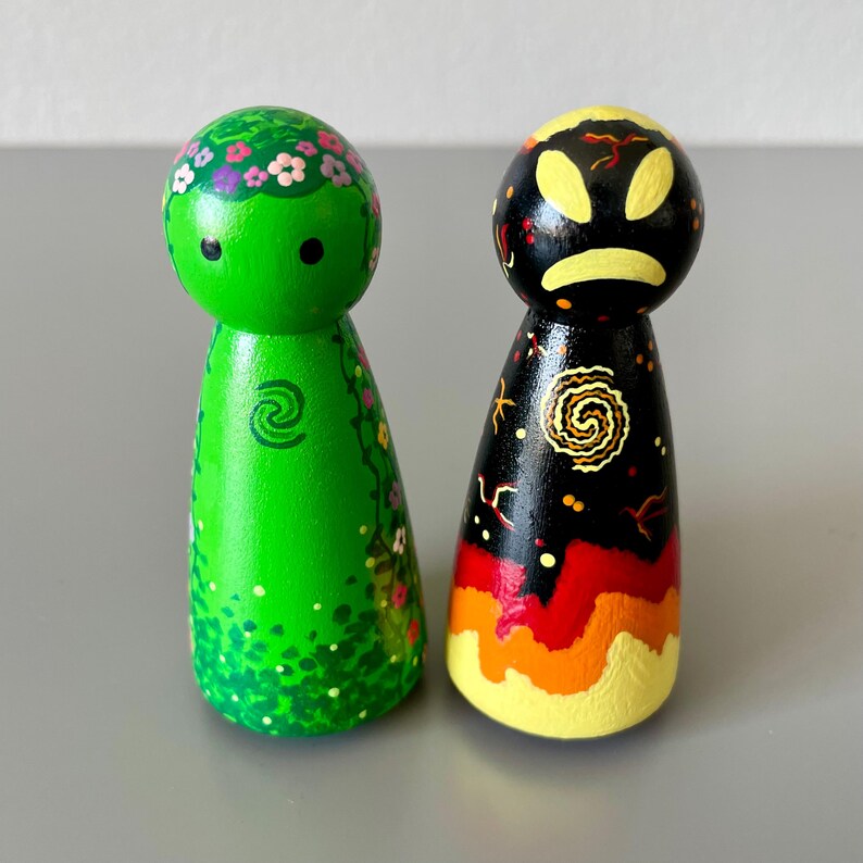 May include: Two hand-painted wooden peg dolls. One is green with floral details and a spiral symbol. The other is black with a yellow face, a spiral design, and a gradient of red, orange, and yellow at the base.