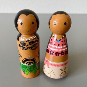 May include: Two hand-painted wooden peg dolls. One doll has black hair and a detailed tattoo design. The other doll has black hair, a pink dress with white polka dots, and a necklace. Both dolls have smiling faces and are on a gray surface.