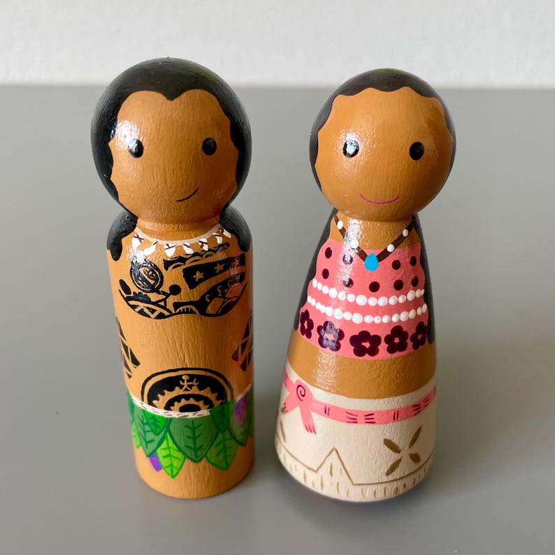 May include: Two hand-painted wooden peg dolls. One doll has black hair and a detailed tattoo design. The other doll has black hair, a pink dress with white polka dots, and a necklace. Both dolls have smiling faces and are on a gray surface.