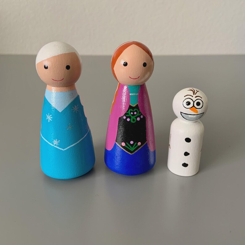 May include: Three wooden peg dolls depicting characters from the animated film. One doll is dressed in a blue dress with snowflake details, another in a pink and purple dress, and the third is a white snowman with a carrot nose.