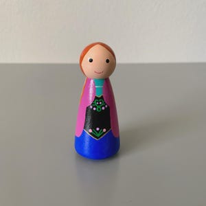 May include: A wooden peg doll with a painted design. The doll has a round head with a smiling face and red hair. The body is painted with a blue skirt, a black bodice with green and pink floral details, and a pink cape.