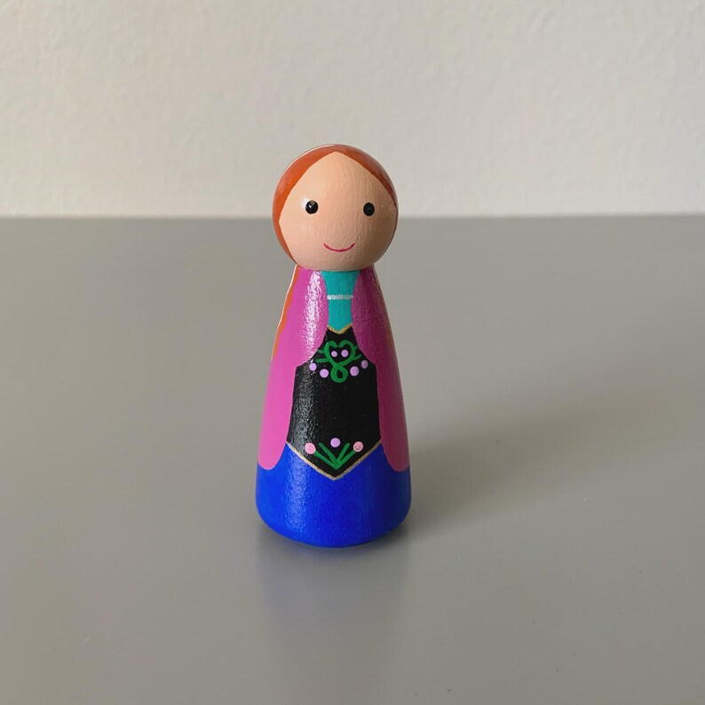 May include: A wooden peg doll with a painted design. The doll has a round head with a smiling face and red hair. The body is painted with a blue skirt, a black bodice with green and pink floral details, and a pink cape.