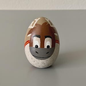 May include: A hand-painted, egg-shaped stone featuring a cartoon donkey face. The donkey has a brown head, white eyes, and a gray muzzle. The egg is tan with white and red accents, creating a whimsical decorative item.