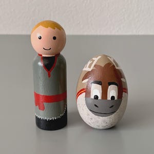May include: Two hand-painted wooden figures: one is a peg doll with a blonde head, gray body, and red sash. The other is an egg-shaped figure with a horse face design in brown, white, and tan. The figures are likely toys or decorative items.