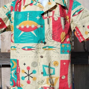 May include: A short-sleeved button-up shirt with a retro-style print featuring geometric shapes, stars, and flying saucers in shades of red, yellow, blue, and green on a cream background.