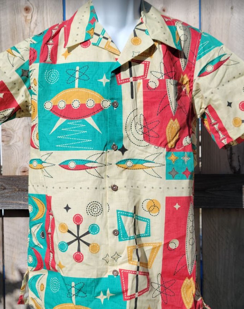 May include: A short-sleeved button-up shirt with a retro-style print featuring geometric shapes, stars, and flying saucers in shades of red, yellow, blue, and green on a cream background.