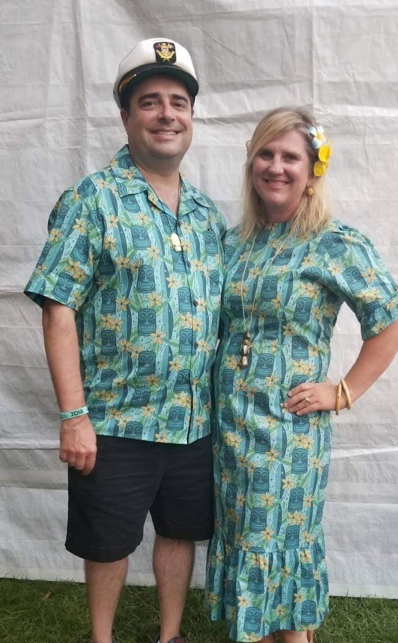 May include: A couple wearing matching blue and green shirts with a tiki design. The man is wearing a white sailor hat. The woman is wearing a flower in her hair.