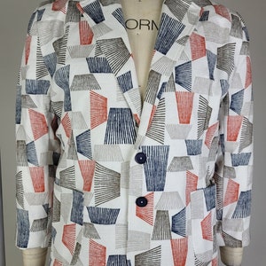 Puede incluir: Una chaqueta blanca con un patrón geométrico en tonos rojos, azules y marrones. La chaqueta tiene un cierre de botón único y dos bolsillos.