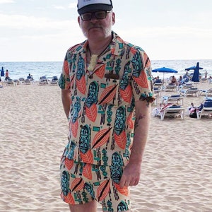May include: A man wearing a beige and orange patterned shirt and matching shorts. The shirt and shorts feature a tiki design with green and blue accents. He is wearing a white trucker hat with the text "Big Beach Tiki Active" on it. The man is standing on a sandy beach with lounge chairs in the background.