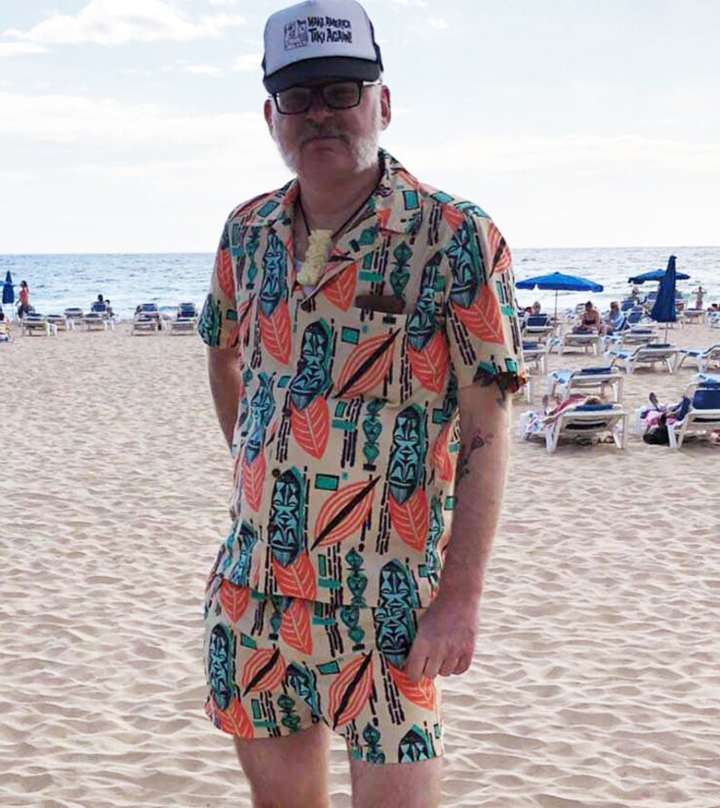 May include: A man wearing a beige and orange patterned shirt and matching shorts. The shirt and shorts feature a tiki design with green and blue accents. He is wearing a white trucker hat with the text "Big Beach Tiki Active" on it. The man is standing on a sandy beach with lounge chairs in the background.