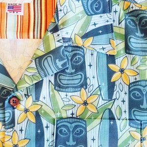 May include: A blue and green Hawaiian shirt with a tiki pattern. The shirt features yellow flowers, tiki faces, and surfboard designs. The shirt has a label that says "Volcano Designs Made in U.S.A."