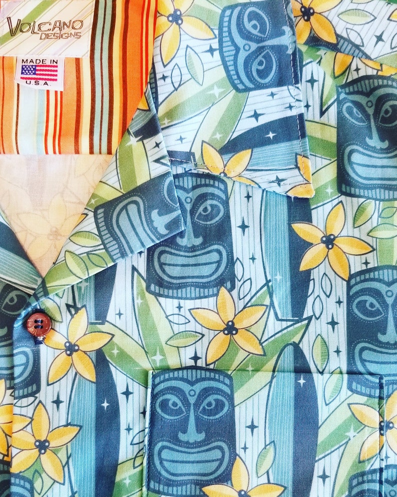 May include: A blue and green Hawaiian shirt with a tiki pattern. The shirt features yellow flowers, tiki faces, and surfboard designs. The shirt has a label that says "Volcano Designs Made in U.S.A."