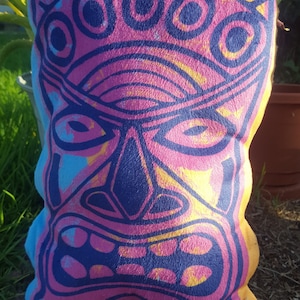 Puede incluir: Una almohada rosa y azul con un diseño de cara estilizada. La cara tiene una boca grande, dos ojos y un tercer ojo por encima de la nariz. La almohada está hecha de tela suave y tiene un borde amarillo.