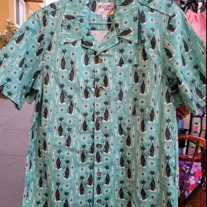 May include: A teal blue short-sleeved shirt with a repeating pattern of black cats and white stars. The shirt has a button-up front and a collar. The label on the shirt reads "Volcano, San Diego".