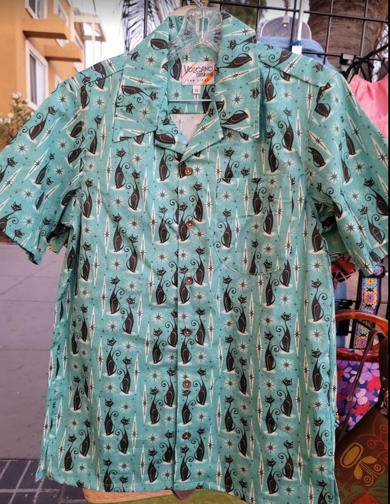 May include: A teal blue short-sleeved shirt with a repeating pattern of black cats and white stars. The shirt has a button-up front and a collar. The label on the shirt reads "Volcano, San Diego".