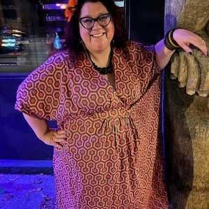 May include: A woman wearing a red and black geometric print dress with a flower in her hair. She is standing next to a large, brown, textured object.