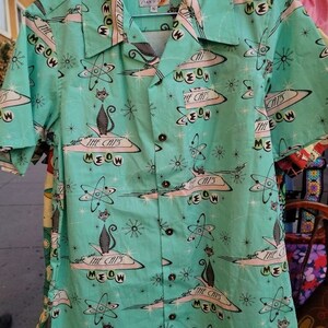 May include: A turquoise blue short-sleeved shirt with a retro print of cats in spaceships and atomic symbols. The shirt has a button-up front and a collar. The text "The Cat's Meow" is repeated throughout the print.