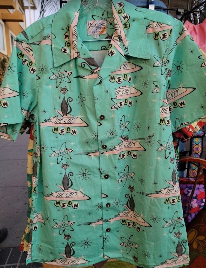 May include: A turquoise blue short-sleeved shirt with a retro print of cats in spaceships and atomic symbols. The shirt has a button-up front and a collar. The text "The Cat's Meow" is repeated throughout the print.