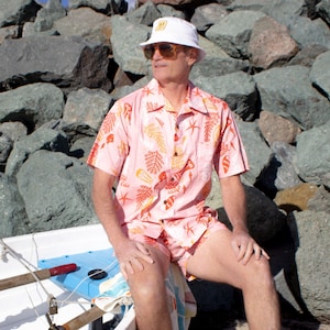 May include: A man wearing a pink and orange Hawaiian shirt with a white bucket hat and sunglasses. He is sitting on a white boat with a blue and red seat.