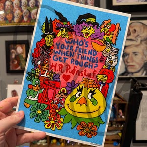 Può includere: Una stampa colorata con personaggi dei cartoni animati e la scritta "WHO'S YOUR FRIEND WHEN THINGS GET ROUGH? H.R. Pufnstuf." L'opera d'arte ha uno sfondo blu con un cuore e fiori e un personaggio giallo sorridente.