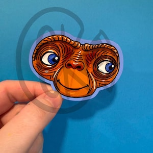 May include: A sticker featuring the face of E.T. the Extra-Terrestrial. The character's head is orange with blue eyes and a smiling mouth. The sticker has a light purple border and is set against a blue background.