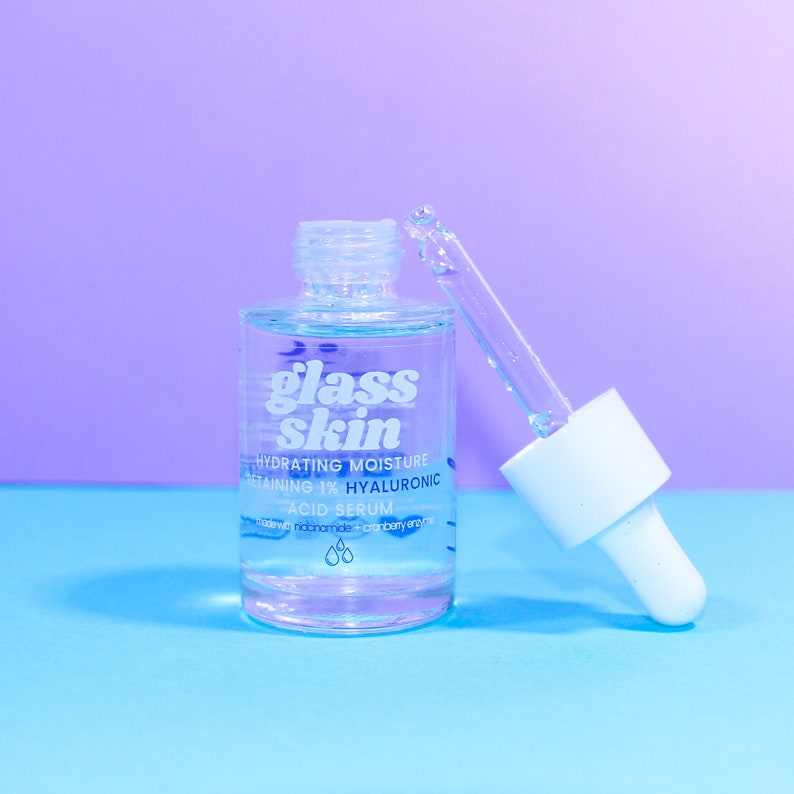 Puede incluir: Una botella de vidrio transparente con un gotero, etiquetada "glass skin hydrating moisture retaining 1% hyaluronic acid serum".