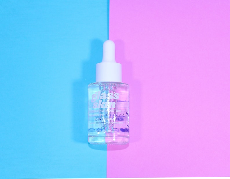 Puede incluir: Una botella de vidrio transparente con un gotero blanco y una etiqueta que dice "glass skin" suero hidratante para la piel con 2% de &aacute;cido hialur&oacute;nico. La botella est&aacute; sobre un fondo azul y rosa.