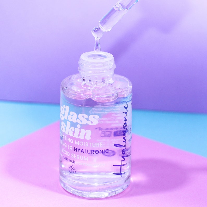 Puede incluir: Una botella de vidrio transparente con un gotero lleno de un l&iacute;quido transparente. La botella est&aacute; etiquetada con "glass skin hydrating moisture 1% hyaluronic acid serum". La botella est&aacute; sobre una superficie rosa y azul.